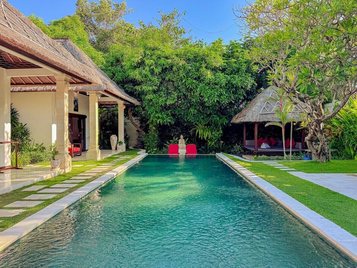 Une piscine rectangulaire dont le centre est d’un bleu limpide, entourée d’une pelouse bien entretenue et bordée, sur la gauche, d’une structure au toit de chaume.
