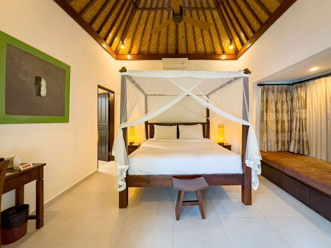 A four-poster bed with a canopy, a wooden bench, a small stool, and a nightstand with a lamp.