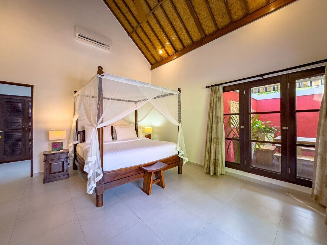 A four-poster bed with a canopy, a small bedside table with a lamp, and a view of a red building through a glass door.