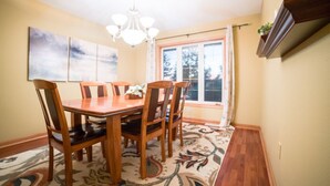 A dining room with a wooden table and chairs, a chandelier, a large window with curtains, and a wall-mounted shelf with a plant. - The Lakeside Retreat (Aberdeen)