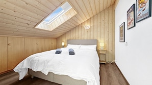 Un dormitorio con techo inclinado, tragaluz, cama con ropa blanca, dos lámparas de noche y cuadros en la pared.