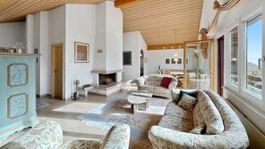 Un salón amplio con chimenea, sofás de estampado floral, techo de madera y ventanas grandes.