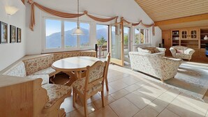 Una habitación amplia con techo de madera, una mesa redonda de comedor, sofás con estampado floral y vistas a montañas a través de ventanas grandes.