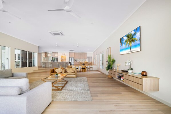 A spacious living area with a sofa, coffee table, and a dining area with chairs. There is a kitchen in the background, a ceiling fan, and a painting of a beach scene on the wall.