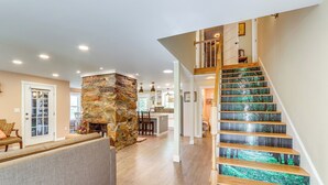 Un vaste espace de vie agrémenté d’une cheminée en pierre, d’un escalier en bois au décor inspiré de la forêt et d’une cuisine située au fond de la pièce.