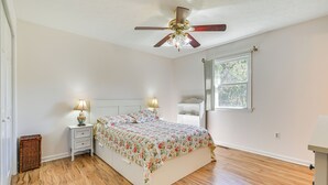 Een slaapkamer met een houten vloer, een bed met bloemenbedrukking, een nachtkastje met lampen, een plafondventilator en een raam met uitzicht op bomen.