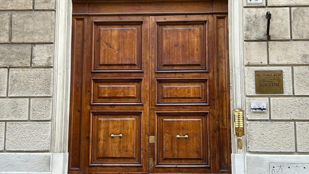 A large wooden double door with a metal decorative arch above it, set in a stone building with a number plaque '19' on the wall.