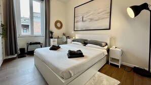 A modern bedroom with a large bed, a desk, a chair, a lamp, and a view of the city through the window.