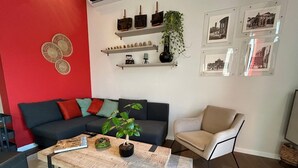 A living room with a red accent wall, a black sofa with colorful cushions, a wooden coffee table with a plant, a beige armchair, and shelves with decorative items and framed pictures.