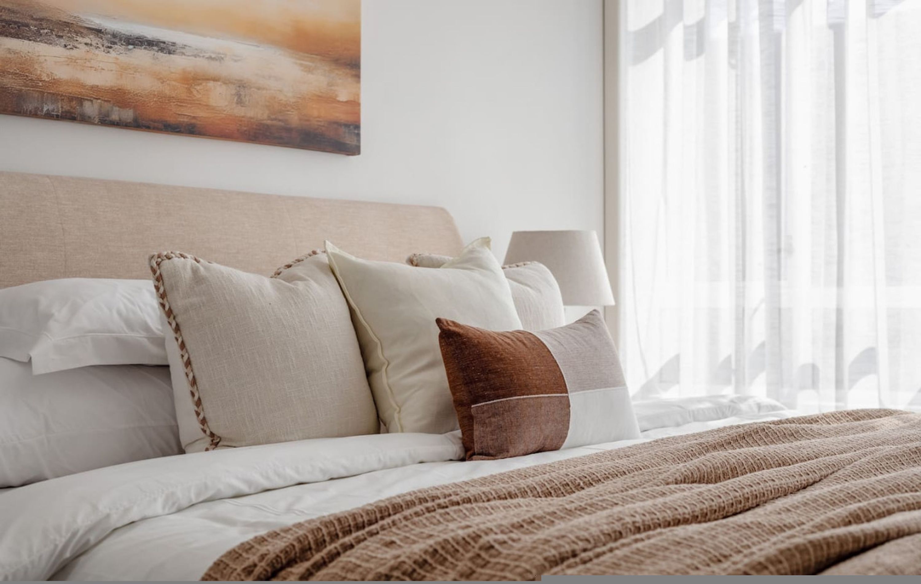 Een net opgemaakt bed met wit en bruin beddengoed, een hoofdbord en een schilderij erboven.