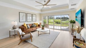 A spacious living room with a large window, a ceiling fan, a sofa, a coffee table, and a TV mounted on the wall.