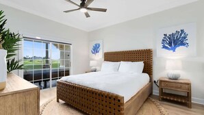 A bedroom with a bed, a ceiling fan, a nightstand, a lamp, a wall-mounted artwork, and a view through a glass door.
