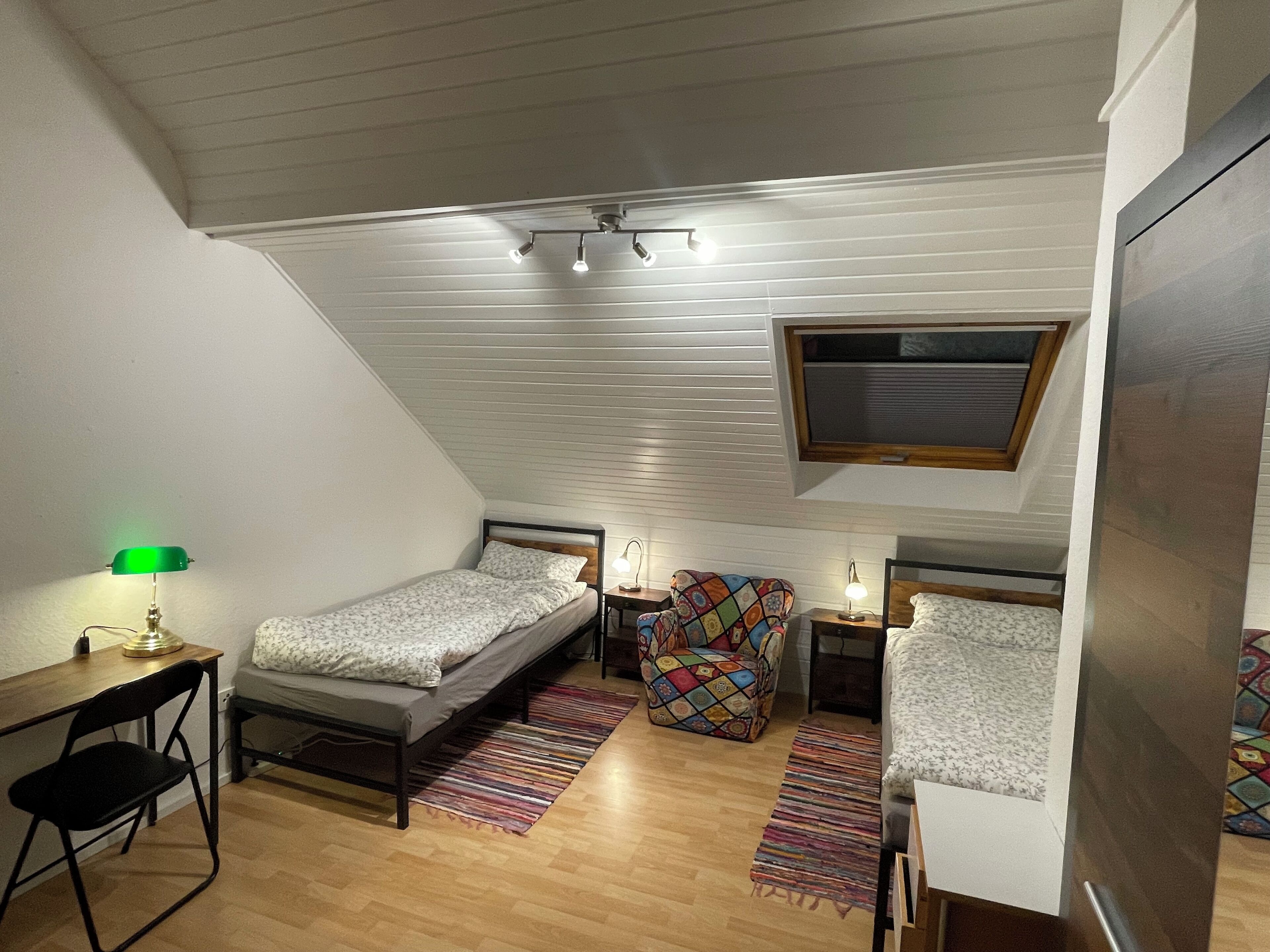 Une petite chambre mansardée, chaleureuse et accueillante, avec un plafond en pente, un bureau avec une lampe, deux lits, une chaise colorée et une petite table.