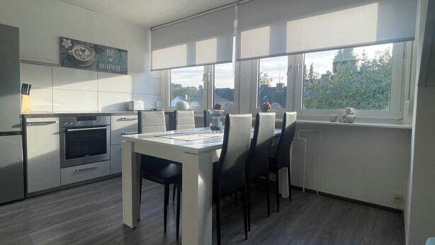 A modern kitchen with white cabinets, a built-in oven, and a dining area with a table and chairs. - Stylish apartment in the middle of the Ruhr area (Bochum)