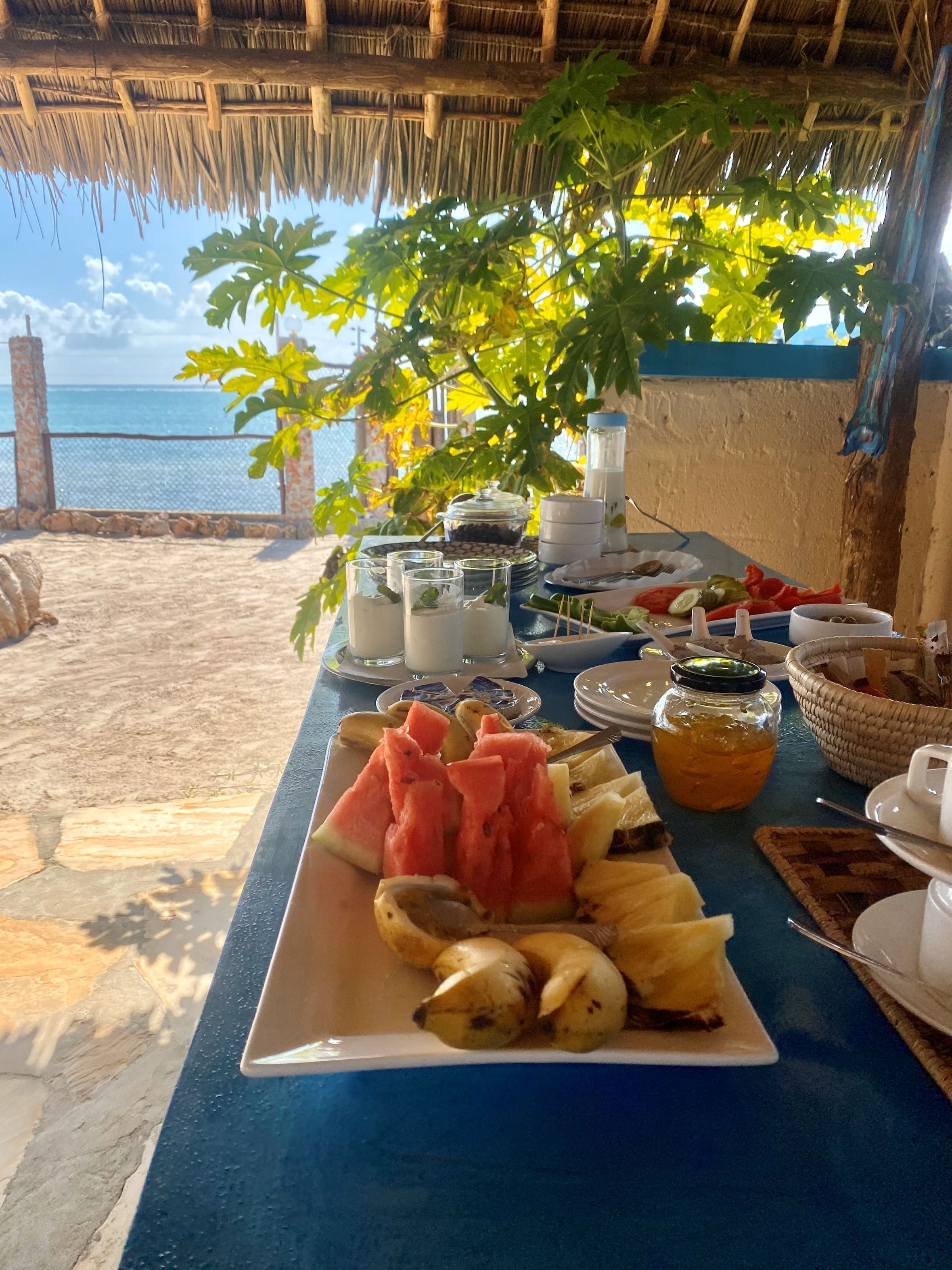A breakfast table with a variety of fruits and beverages, overlooking a beach with a clear view of the sea.