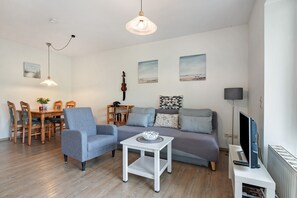 A living room with a sofa, armchair, dining table, and a TV stand. - Apartment 'Wurster Nordseeküste' with Balcony and Wi-Fi (Wurster Nordseeküste)