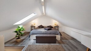 Une chambre avec un plafond en pente, un puits de lumière, un lit avec des oreillers, des lampes de chevet, une plante en pot et un banc.
