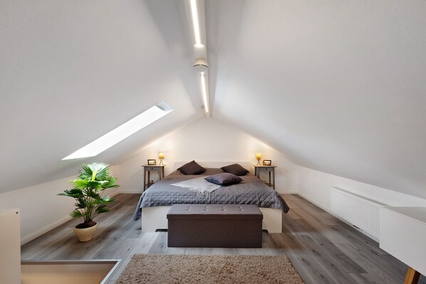 Un dormitorio con techo inclinado, tragaluz, cama con almohadas, lámparas de noche, planta en maceta y un banco.