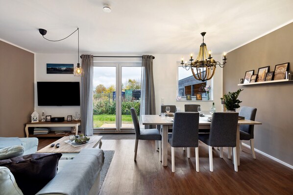 A modern living room with a flat-screen TV, a dining area with a table and chairs, and a large window with a view of the outdoors. - Holiday Home 'Küsten-Bude' with Private Terrace, Garden, and Wi-Fi (Elmenhorst)