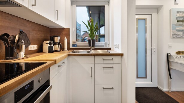 Une cuisine moderne avec des armoires blanches, un plan de travail en bois, un four encastré, une machine à café et une fenêtre donnant sur des immeubles.