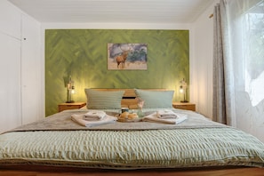 Un letto rifatto con cura, una parete verde, un quadro che raffigura un cervo e un vassoio con pane e una tazza.