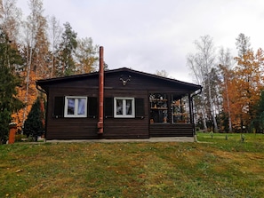 A wooden cabin with a chimney, two windows with shutters, and a porch with a railing. - Holiday Home 'Sonnenpark 44' with Lake View, Private Terrace and Wi-Fi (Roding)