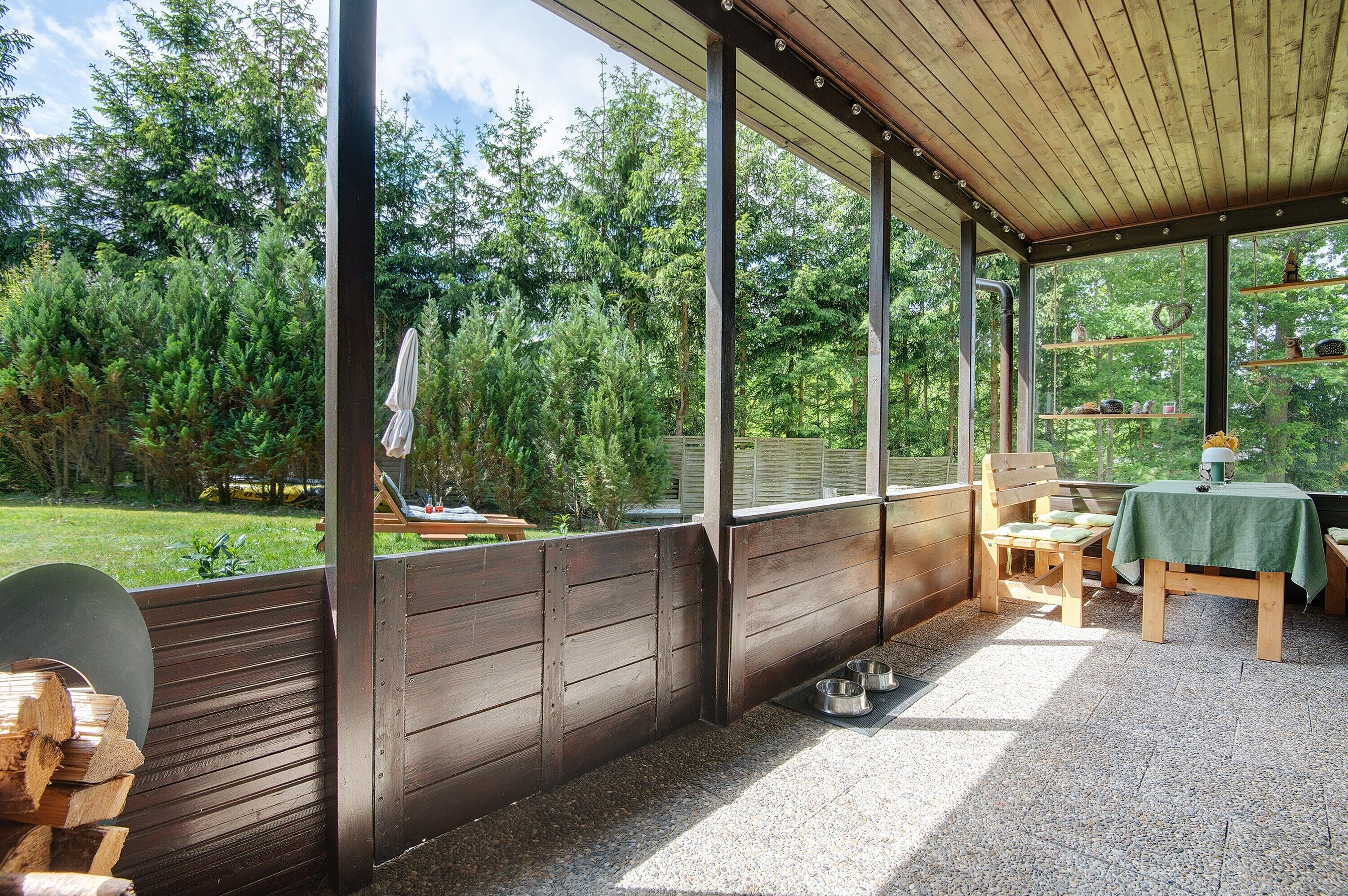 Een overdekt buitengebied met een houten bank, een tafel met een groene tafelkleed en voerbakken voor huisdieren op de vloer.