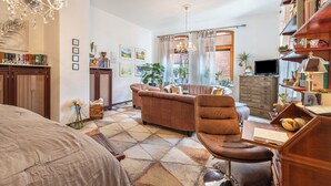 Un salon bien éclairé, avec un canapé, un fauteuil et un téléviseur. On y trouve également des étagères à livres, un lustre et une fenêtre avec des rideaux.