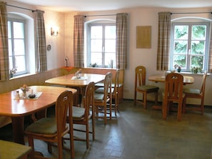 Een eetgedeelte met houten tafels en stoelen, grote ramen met gordijnen en een betegelde vloer.