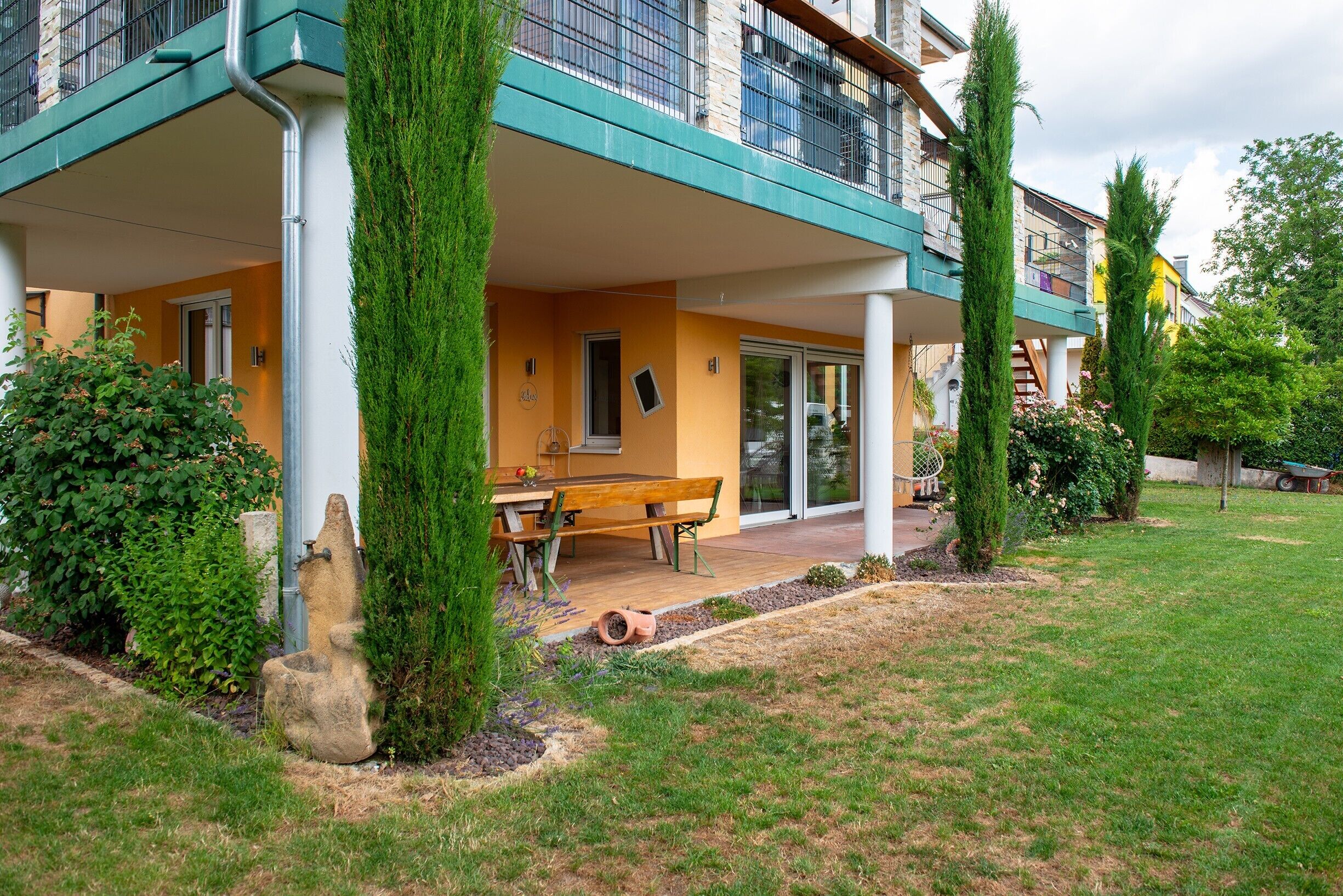 Un edificio moderno con un patio cubierto, un comedor con una mesa y bancos, y un jardín bien cuidado con árboles altos y arbustos.
