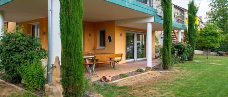 Un edificio moderno con un patio cubierto, un comedor con una mesa y bancos, y un jardín bien cuidado con árboles altos y arbustos.
