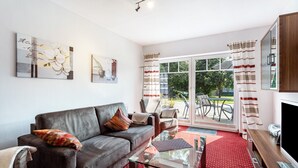 Un salon comprenant un canapé, un fauteuil, une table basse en verre et un téléviseur. Une porte-fenêtre coulissante donne accès à un espace extérieur aménagé avec une table et des chaises.