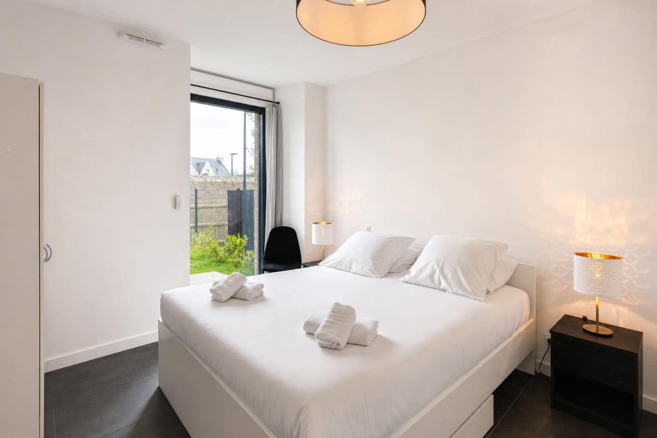 A modern hotel room with a large bed, two bedside tables with lamps, a view of greenery outside, and a door to the left.