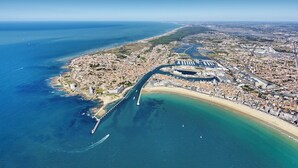 Vue aérienne d’une ville côtière, avec un port de plaisance bien visible, une vaste plage de sable et une zone urbaine dense, caractérisée par des bâtiments et des routes.