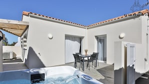 Un jacuzzi en un espacio exterior techado con una mesa y sillas, una puerta corrediza y una pared blanca con techo de azulejos.