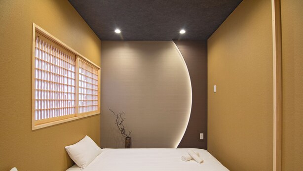 A hotel room with a bed, a window with blinds, a wall-mounted vase with branches, and recessed ceiling lights. - Travel time Setsugekka (Osaka)