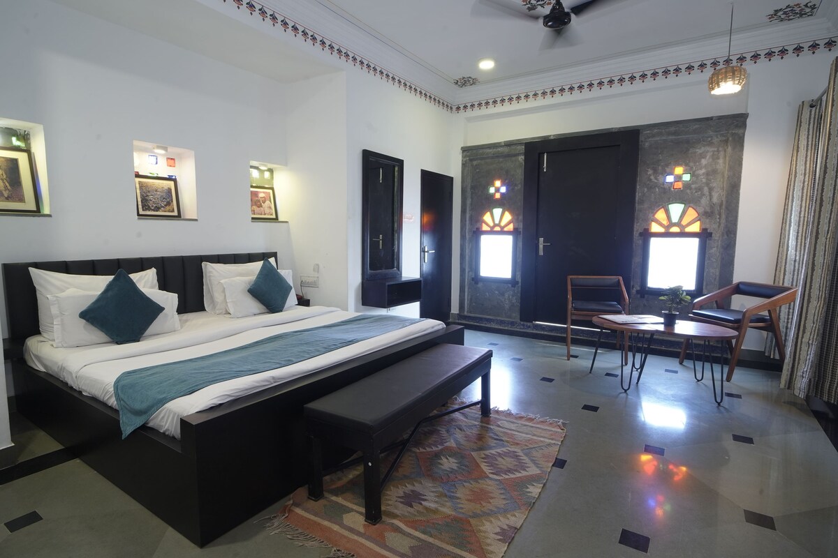 A modern hotel room with a large bed, a bench, a small table, and a view of the outside.