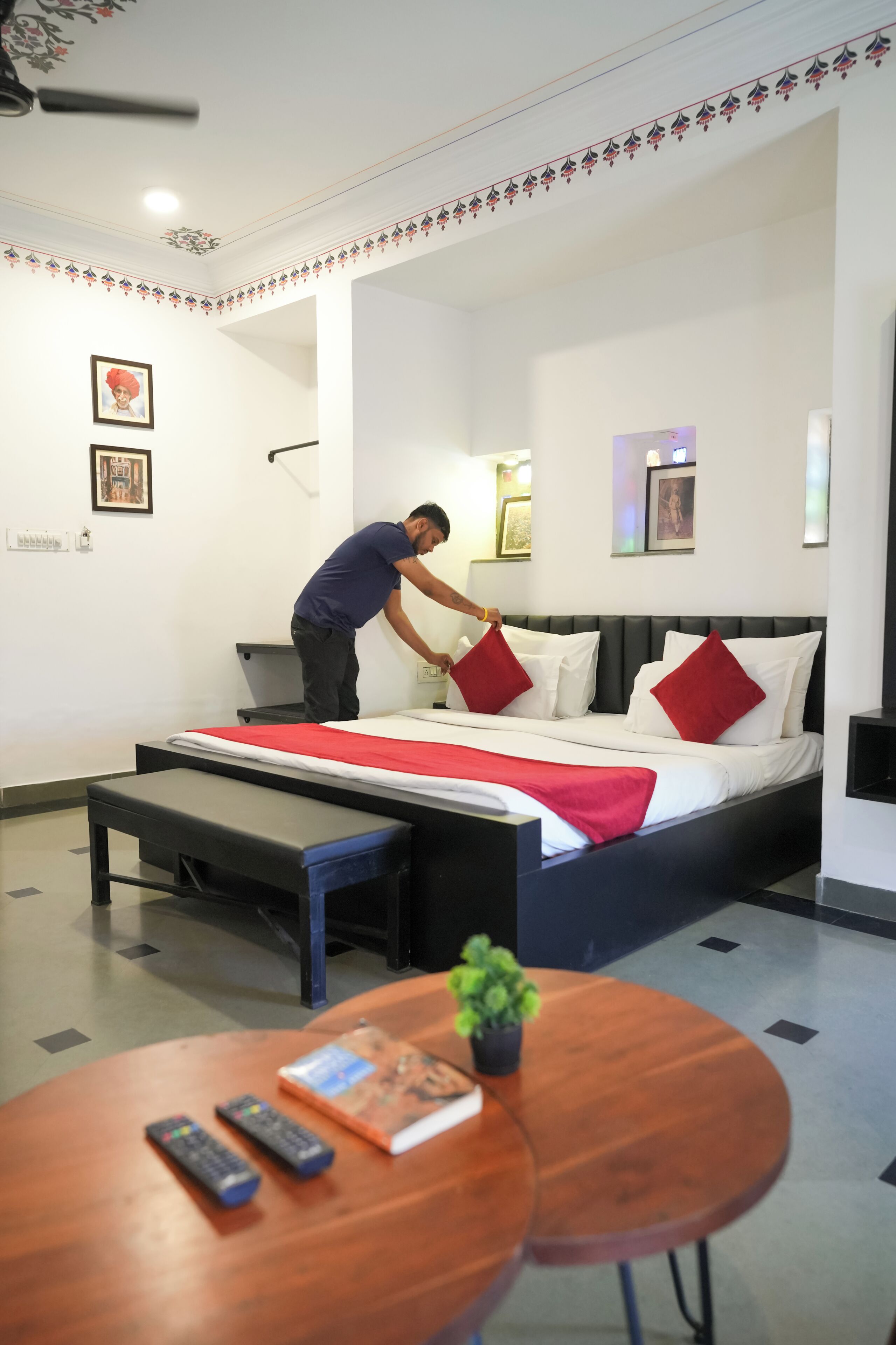 Une chambre d’hôtel avec un lit, des tables de chevet, une petite table avec un livre et des télécommandes, et un ventilateur de plafond.