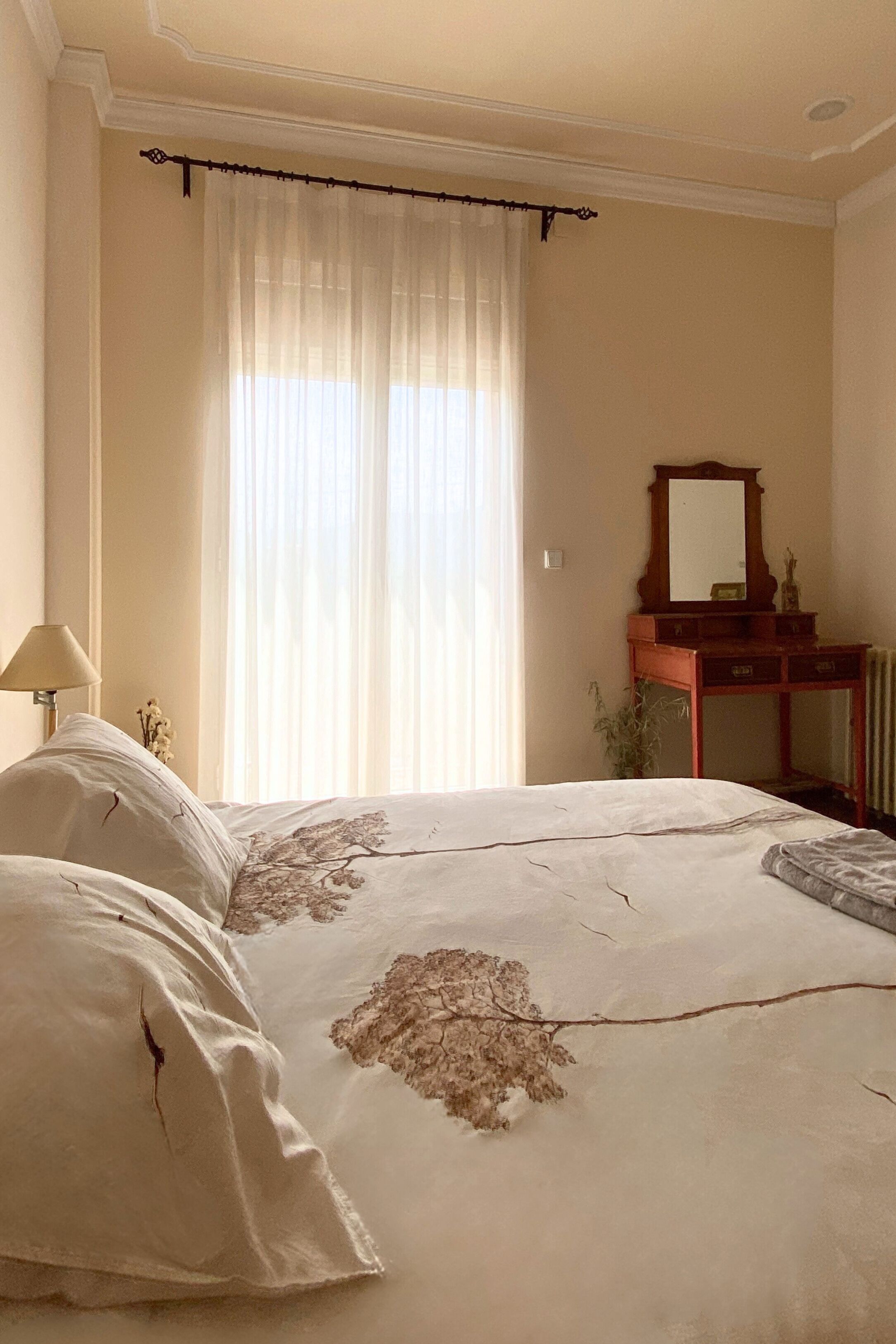 A bedroom with a bed, a dresser, a mirror, and a window with sheer curtains.