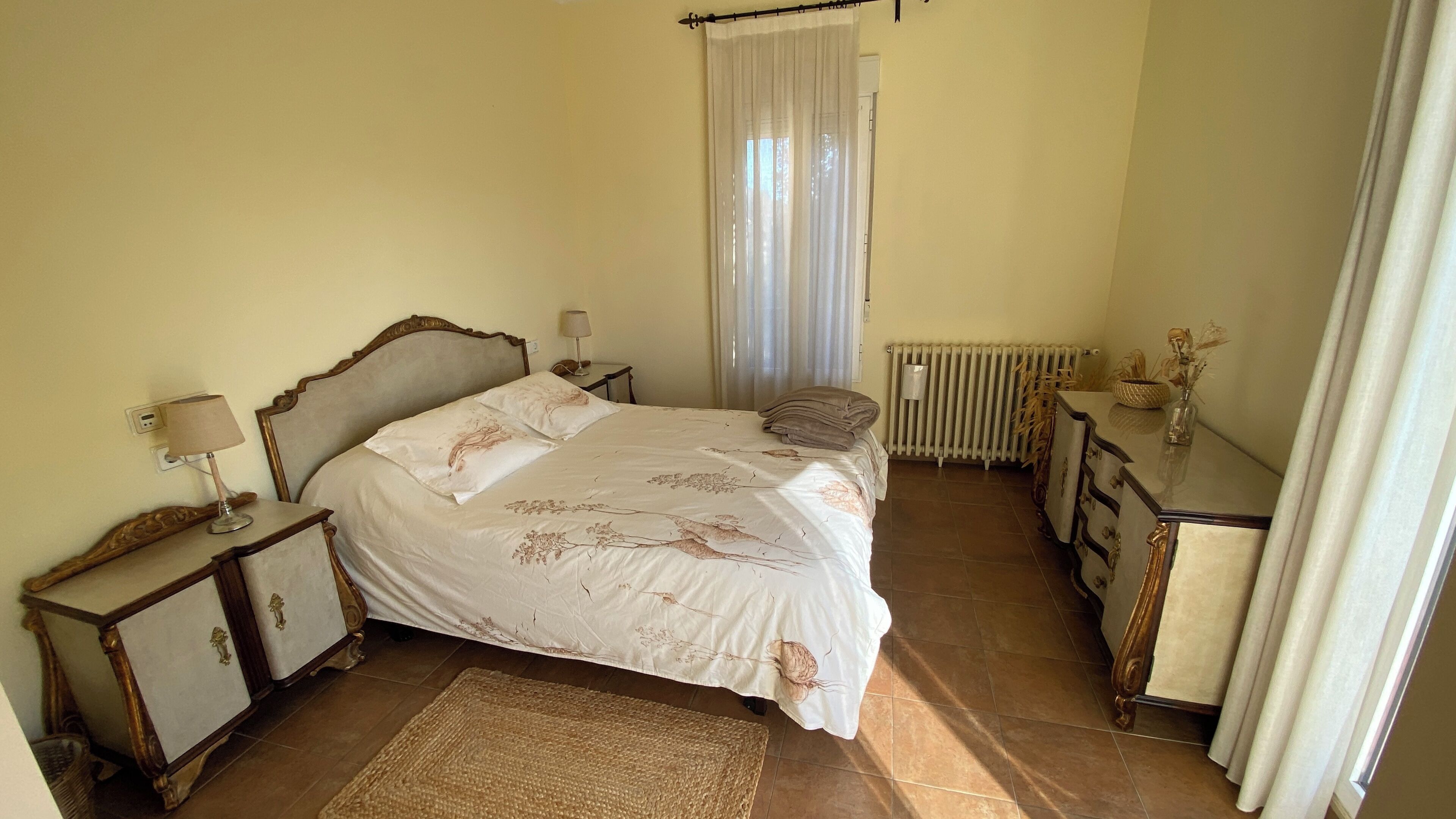 A bedroom with a bed, bedside tables, a dresser, a radiator, and a window with curtains.