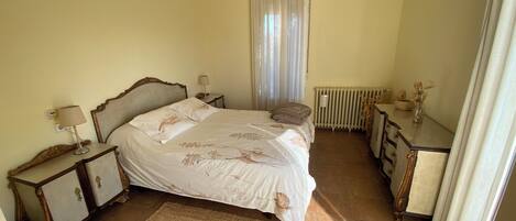 A bedroom with a bed, bedside tables, a dresser, a radiator, and a window with curtains.