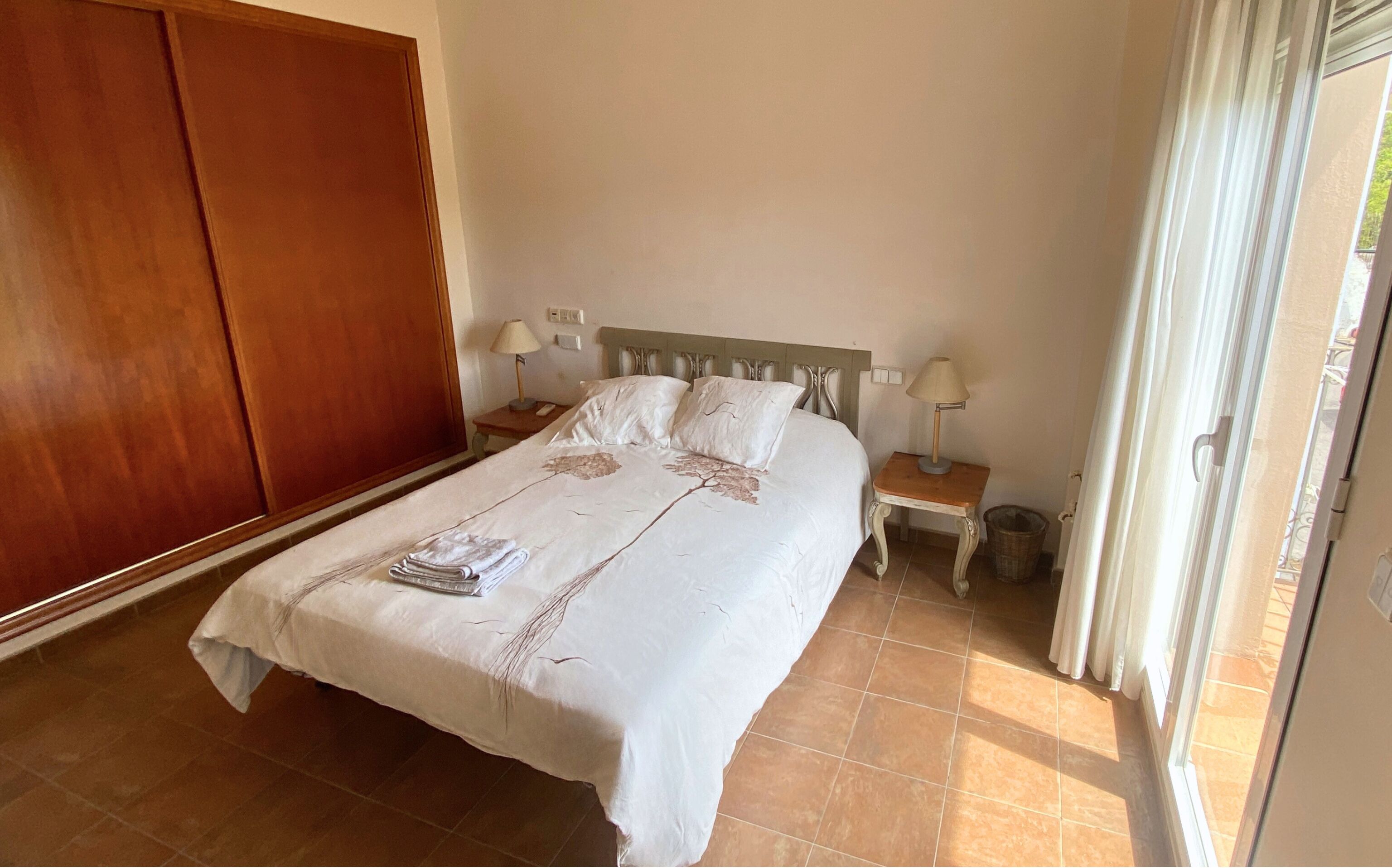 A neatly made bed with a white bedspread and pillows, a wooden wardrobe, two bedside tables with lamps, and a view of the outdoors through a window with curtains.
