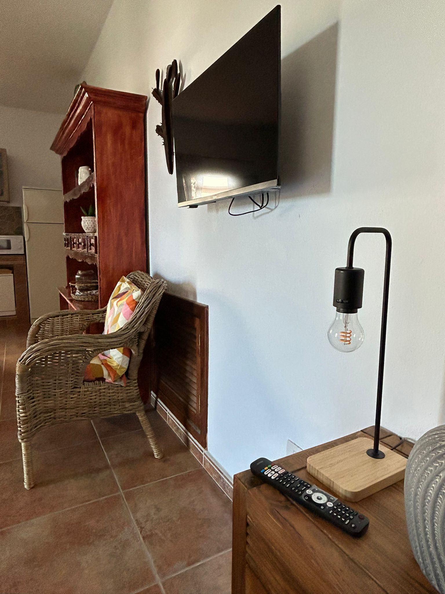 Una sala de estar con un televisor de pantalla plana montado en la pared, un sillón de mimbre con un cojín colorido, una mesita de madera con un control remoto y una lámpara de pie con pantalla de vidrio.