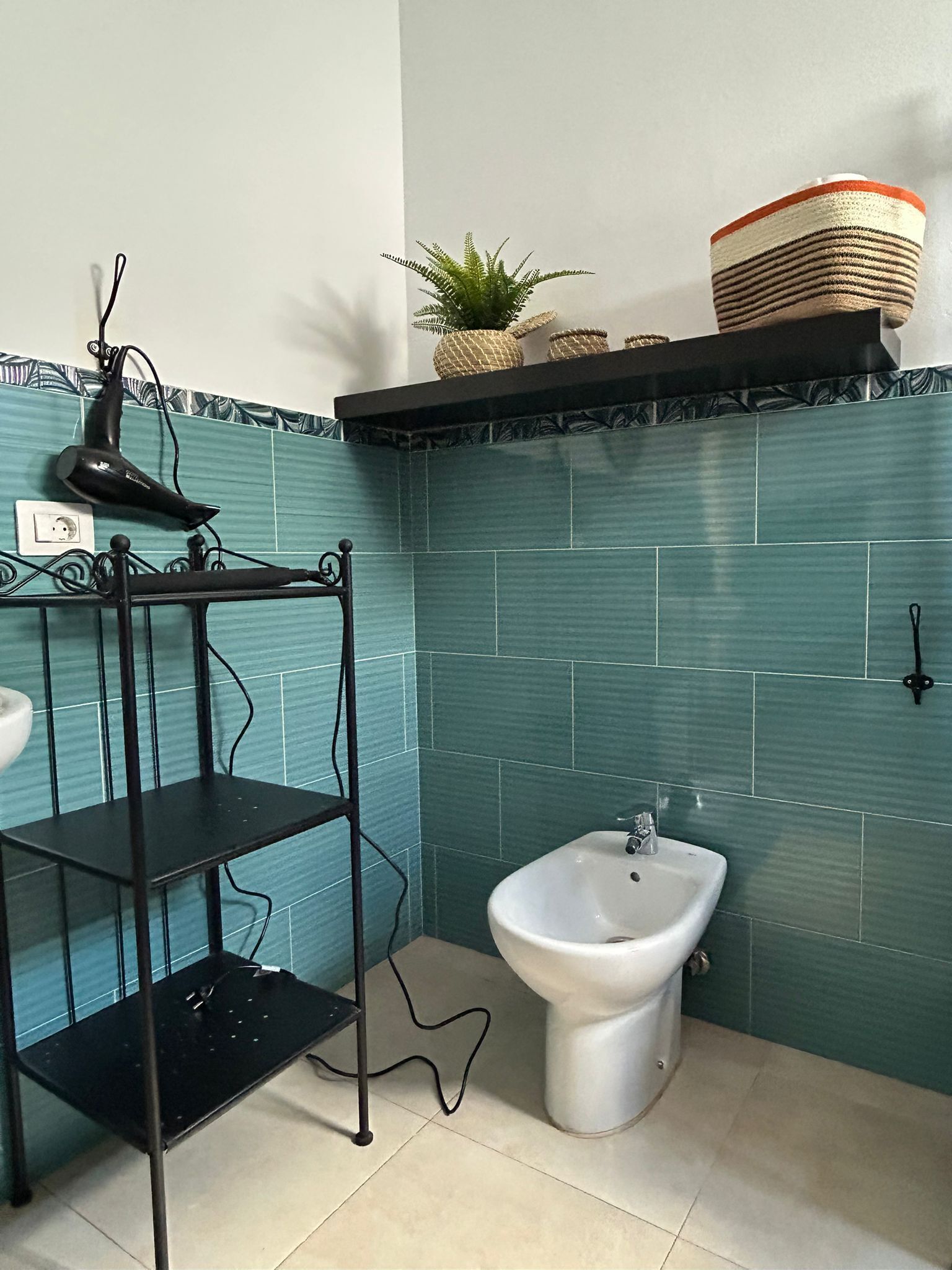 A bathroom with teal tiles, a white sink, a bidet, a hairdryer on a stand, and a shelf with a plant and basket.