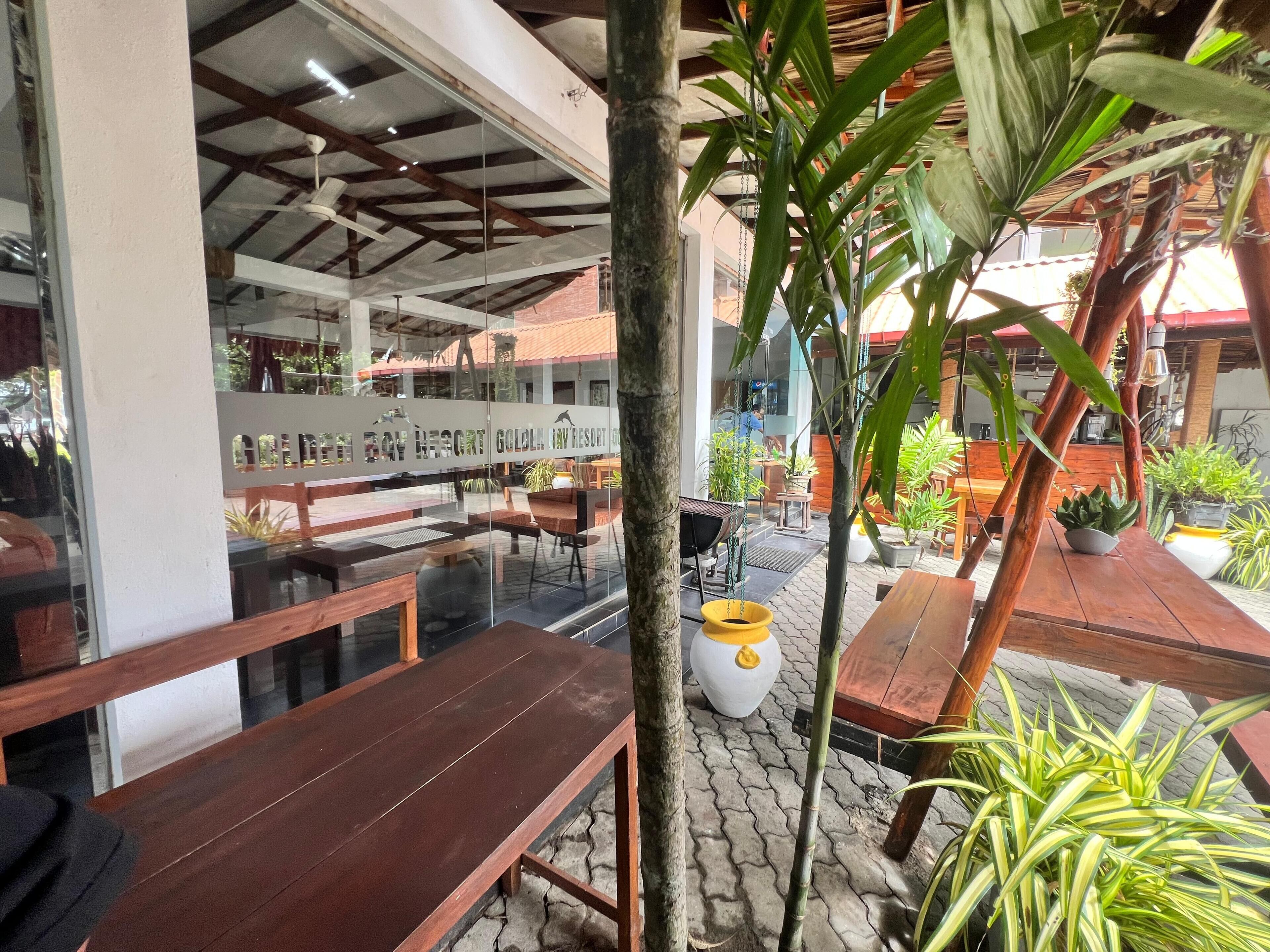 An outdoor seating area with wooden tables and chairs, surrounded by potted plants and a glass wall with the name "Golden Bay Resort".