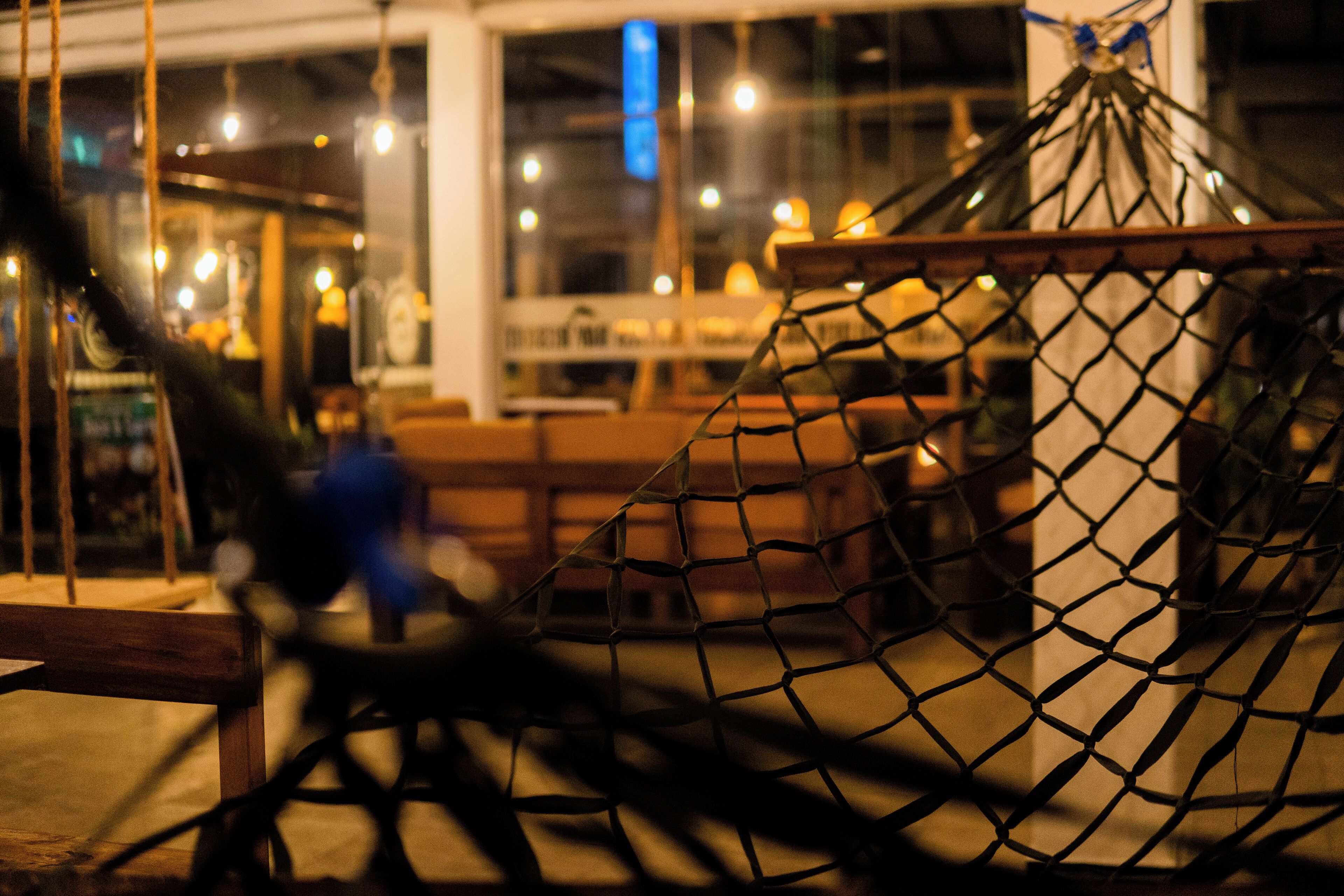 A hammock suspended between wooden beams in a well-lit indoor space with tables and chairs in the background.