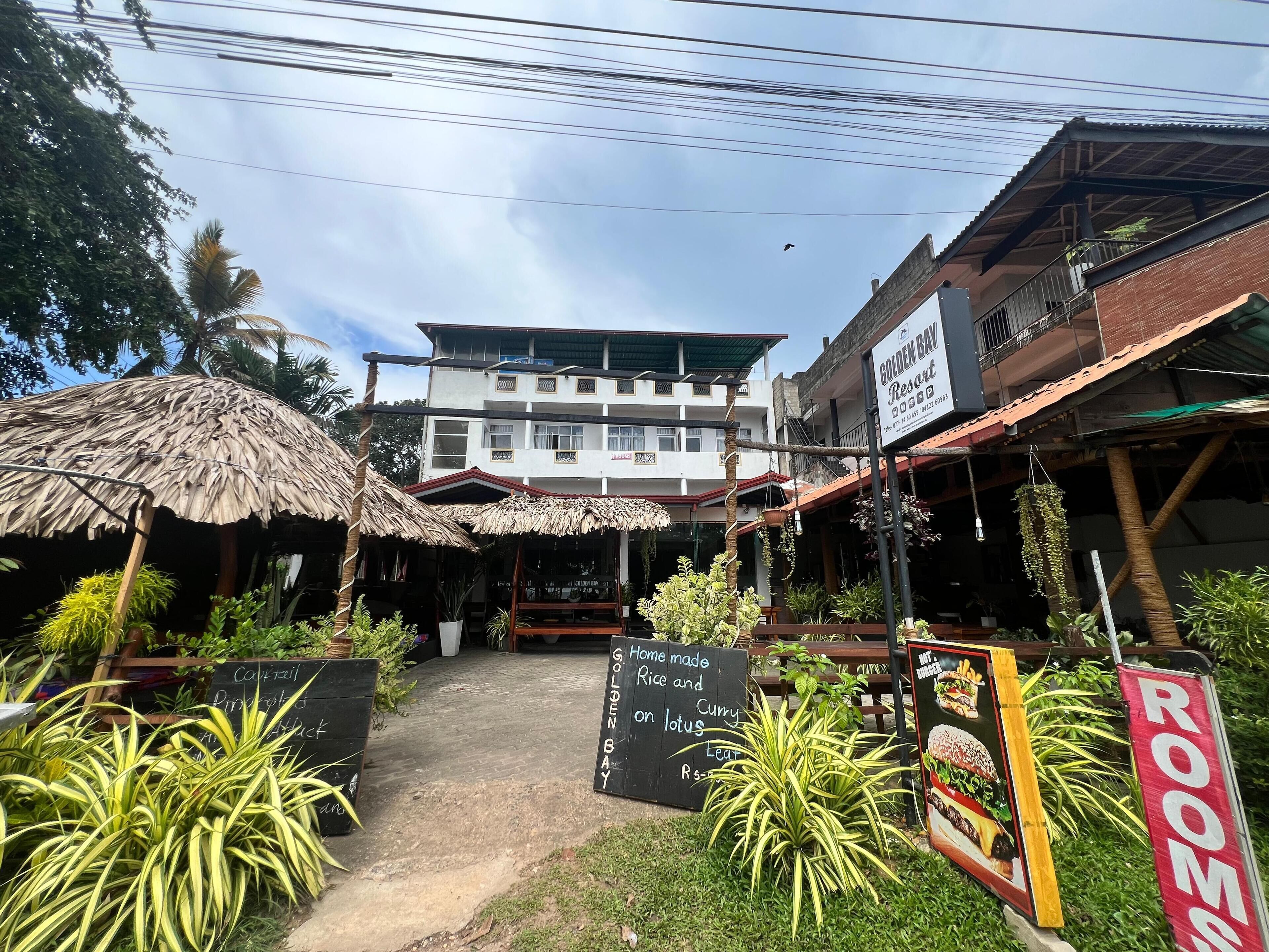 Un resort con una estructura de techo de paja, un letrero que indica habitaciones y un cartel anunciando un restaurante de cocina local.