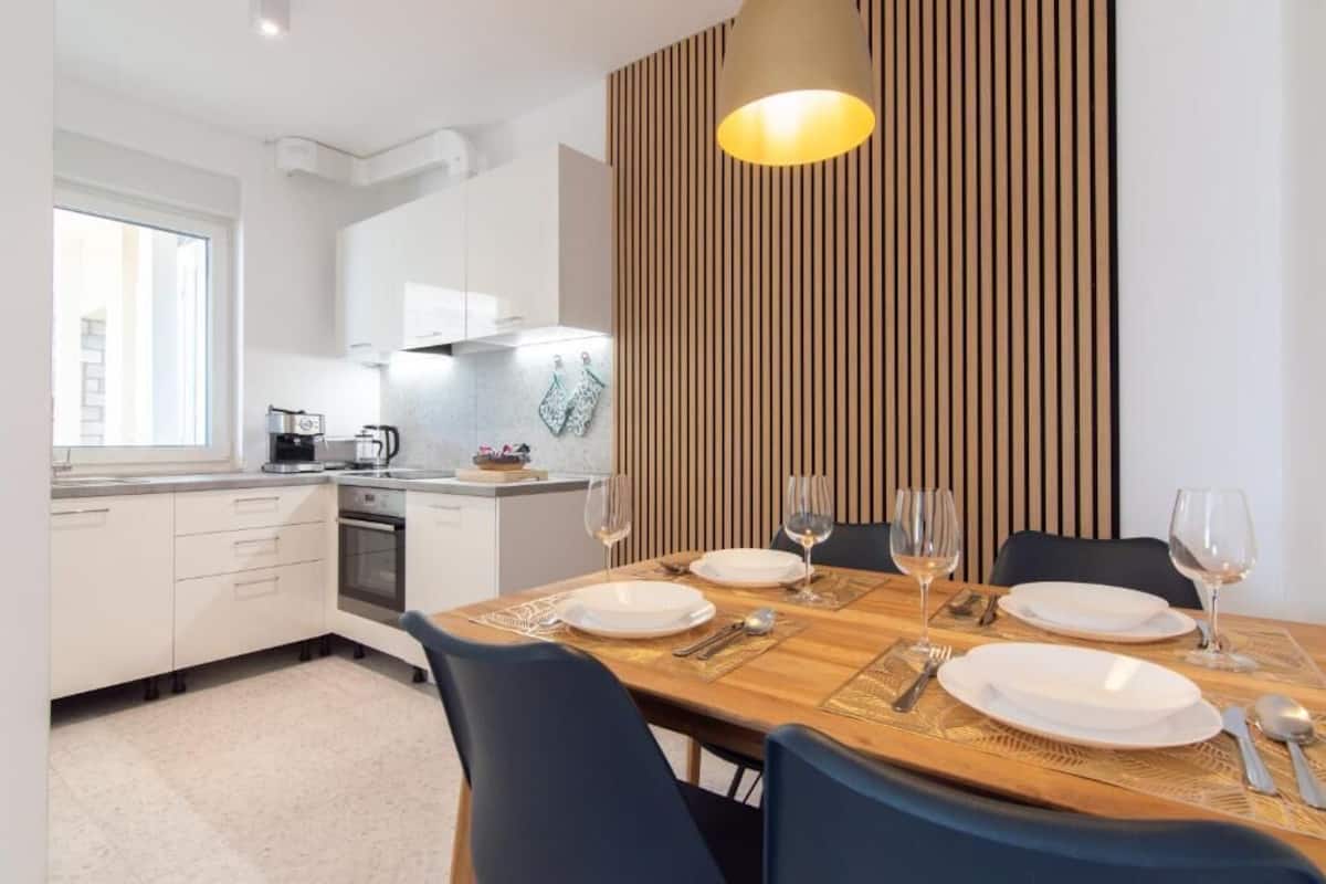 Una cocina moderna con armarios blancos, una mesa de comedor de madera para cuatro personas y un panel de madera vertical en la pared.