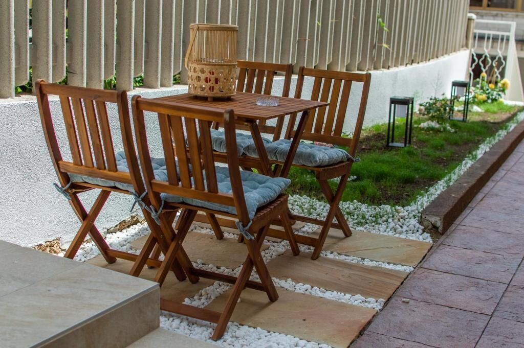 Un'area salotto esterna in legno con tavolo e sedie, un cesto di vimini e un piccolo giardino con ghiaia bianca e piante verdi.