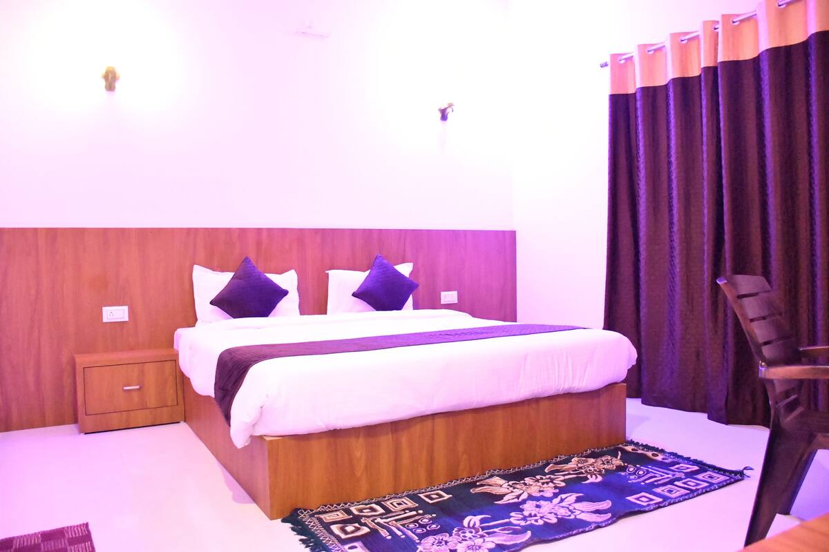 Un dormitorio con cabecera de madera, cama con ropa blanca y almohadas moradas, una mesita de noche, una silla y una alfombra estampada.